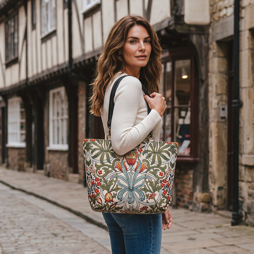A woman wearing a cream jumper and blue jeans holding a large Arts & Crafts style shoulder bag with a floral pattern in green, orange, and blue, walking down a cobblestone street in York. By Umpie Handbags