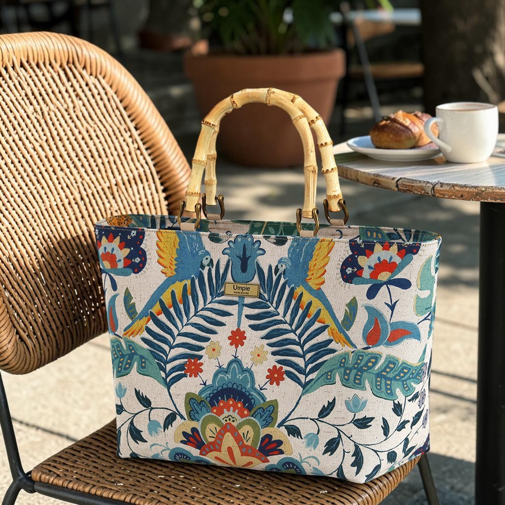 Front view of a floral linen handbag with bamboo handles standing upright on a chair, showcasing the full botanical floral print and the warm natural curve of the bamboo handles, by Umpie Handbags