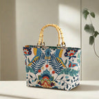 Front view of a floral linen handbag with bamboo handles standing upright on a chair, showcasing the full botanical floral print and the warm natural curve of the bamboo handles, by Umpie Handbags