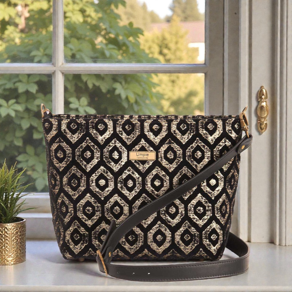  Luxurious gold metallic fabric crossbody bag with sleek black leather strap, featuring firm structured construction and protective metal feet on base, front view showcasing shimmering surface and zip-top closure by Umpie Handbags