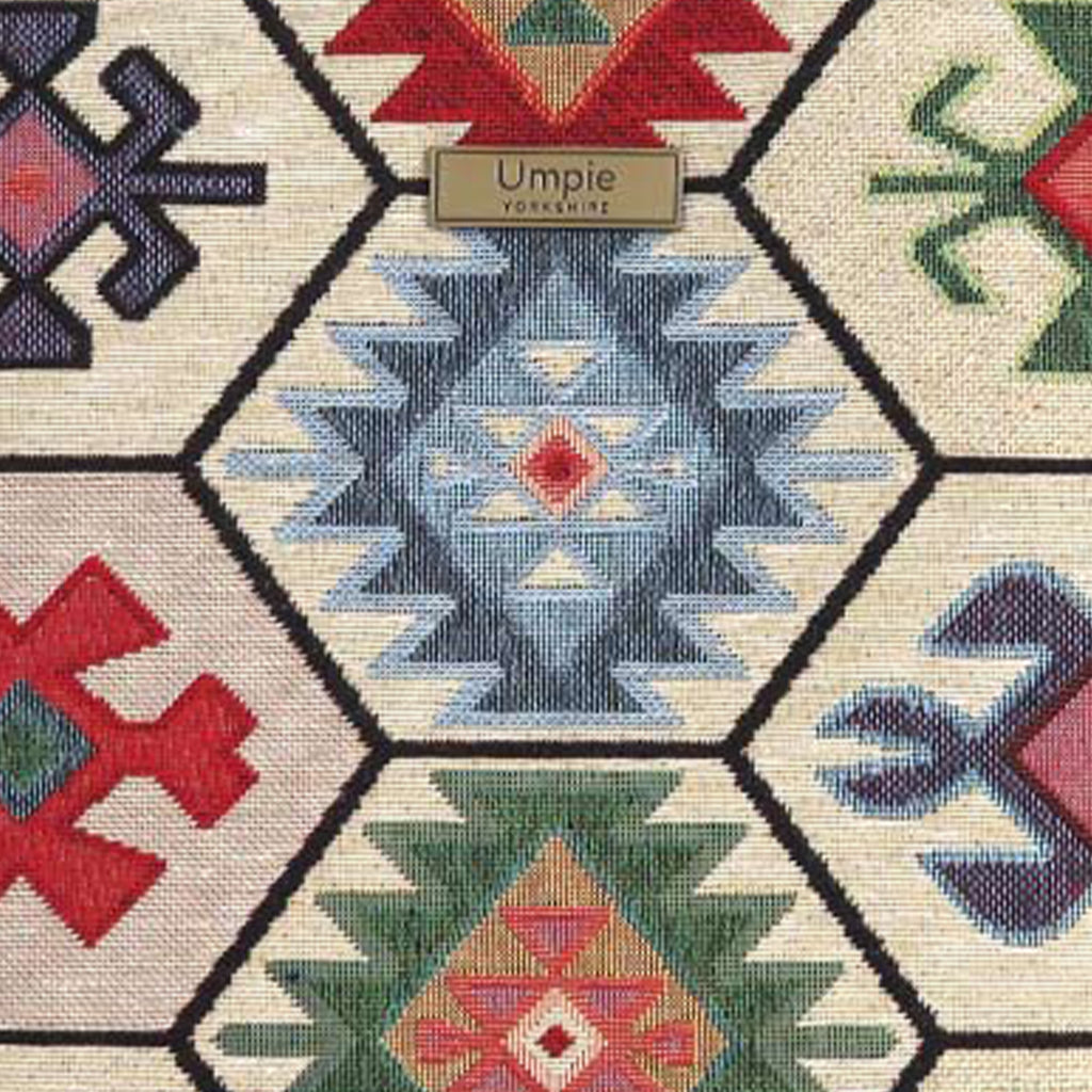 Close-up of the Hexagon Handbag's kilim-inspired tapestry fabric showing the intricate woven texture of individual geometric motifs in blue, red, green and navy on a cream ground, with the Umpie Yorkshire brass badge, by Umpie Handbags