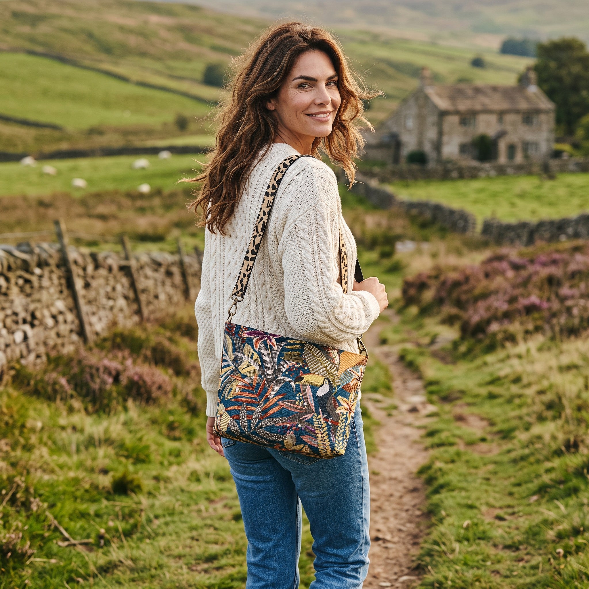  Woman wearing the Toucan Tapestry Crossbody Bag on a Yorkshire moorland path, cable knit jumper, heather in bloom, dry stone walls, leopard print strap visible, by Umpie Handbags