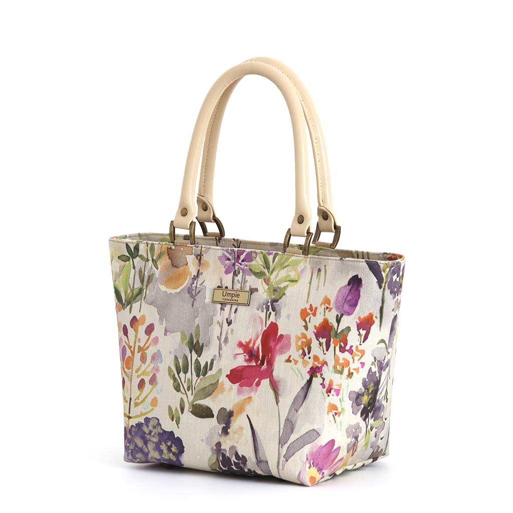Front angled view of Wildflower Handbag on white background, painterly watercolour wildflower print with cream leather handles and antique bronze hardware, by Umpie Handbags
