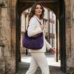 A model in a cream trouser suit walks through a stone archway in York while carrying a large, slouchy purple velvet hobo bag with a leopard print leather strap by Umpie Handbags