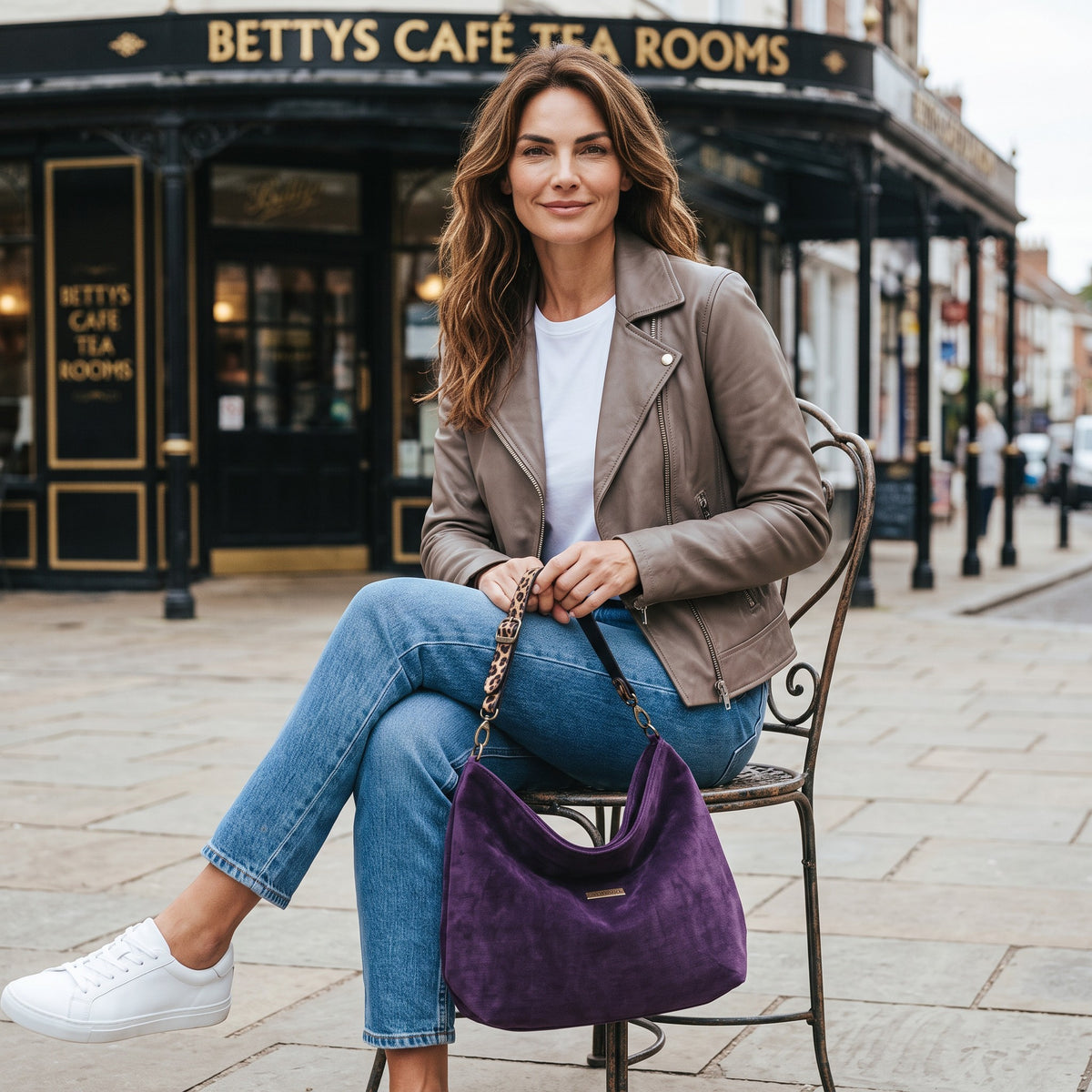 A woman seated on a street in York in front of Bettys tea rooms, carrying a purple velvet hobo bag with a leopard leather strap, by Umpie Handags
