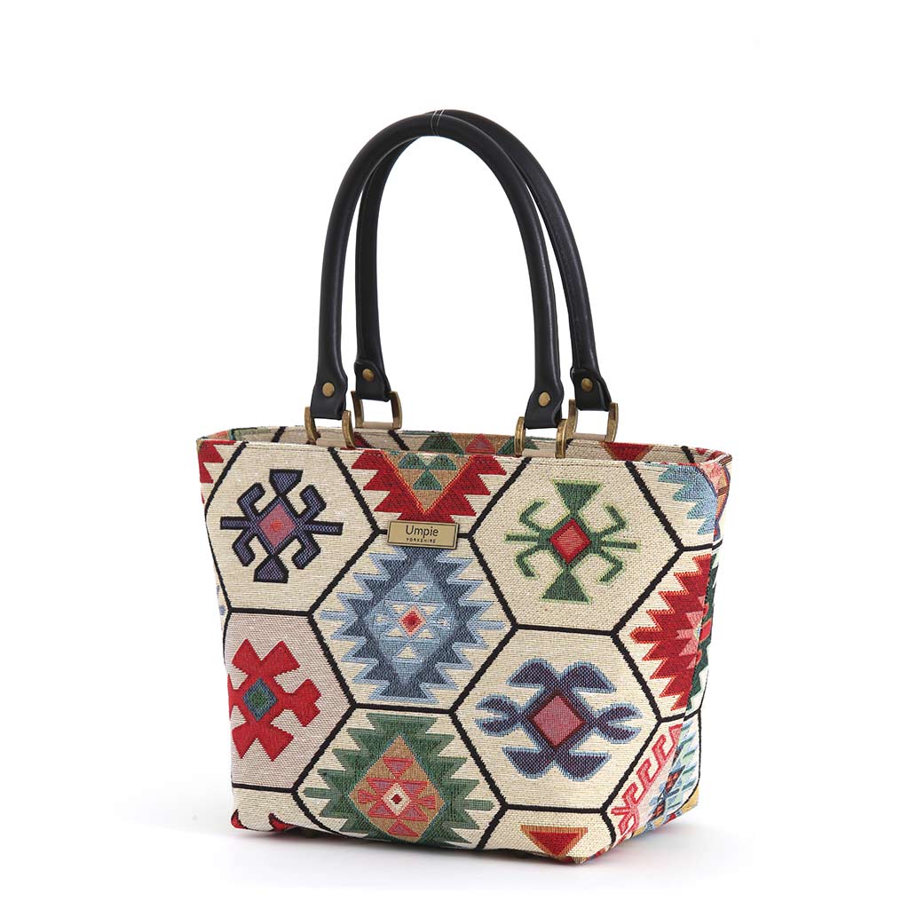 Front view of the Hexagon Handbag in multicoloured kilim-inspired tapestry fabric, each hexagon panel featuring a unique geometric motif in red, blue, green and navy, with black leather handles and antique brass fittings, by Umpie Handbags