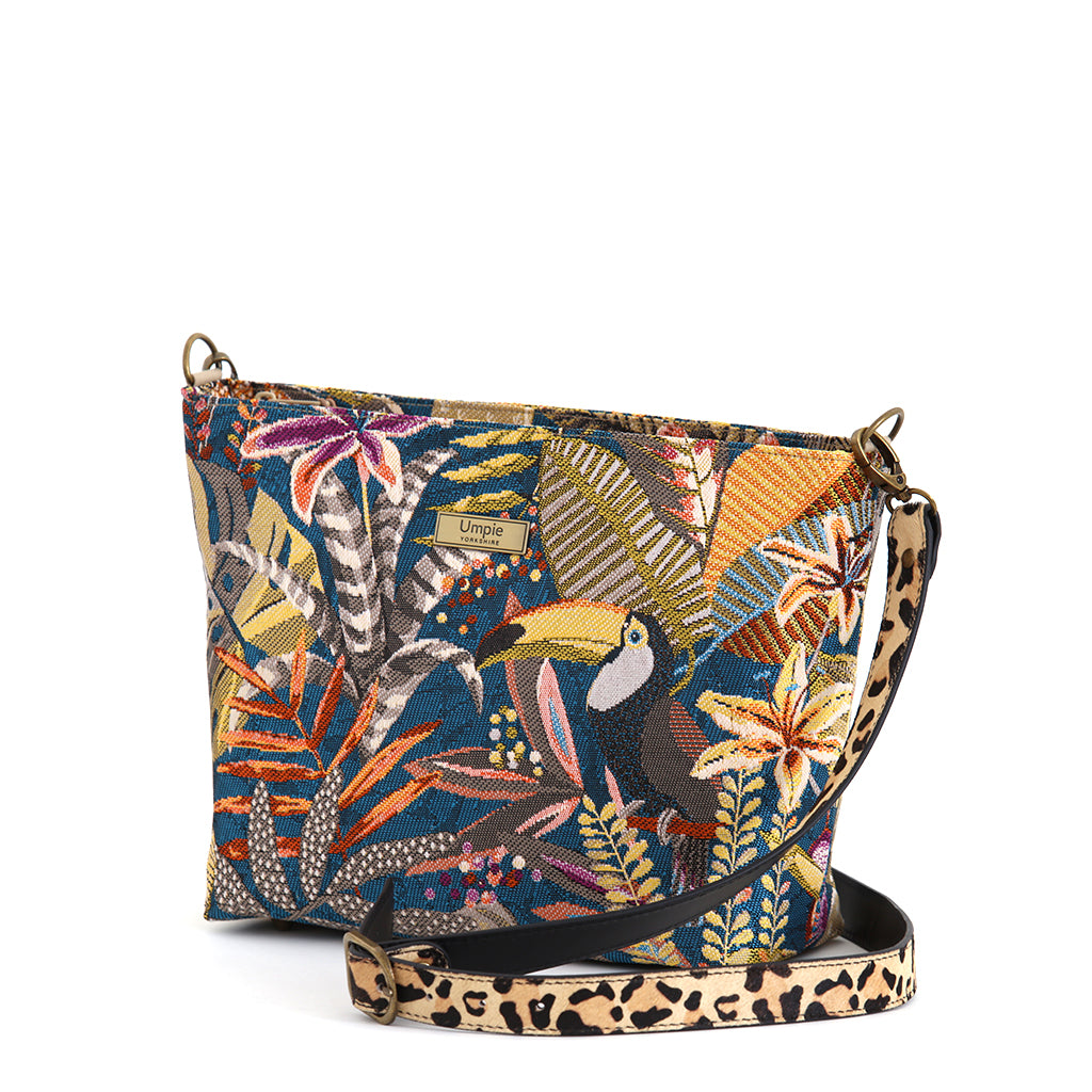 Toucan Tapestry Crossbody Bag front view, richly woven teal tapestry fabric with toucan, jewel-toned botanical foliage in warm golds and burnt orange, leopard print leather strap, antique gold hardware, by Umpie Handbags