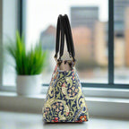 William Morris Handbag with a floral pattern and black leather handles, on a surface near a window with a plant in the background