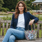 Model seated on a park bench in a Yorkshire garden carrying the William Morris Golden Lily handbag in rich navy tapestry with botanical lily blooms in sage green, blue and coral, with premium black leather handles, handmade in Yorkshire by Umpie Handbags