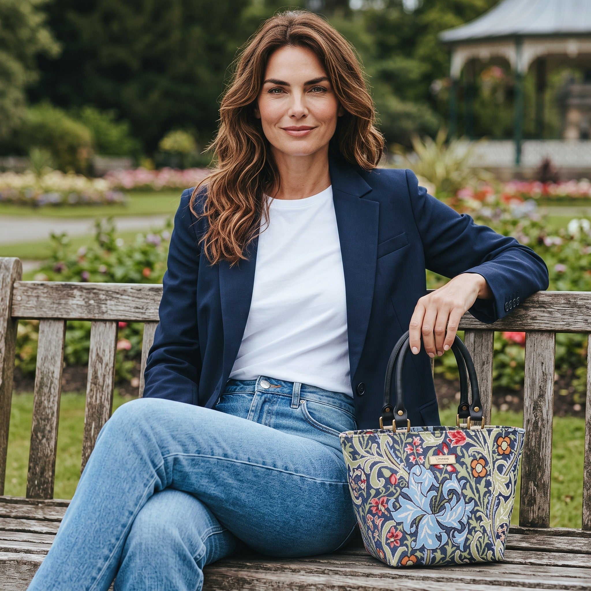 Model seated on a park bench in a Yorkshire garden carrying the William Morris Golden Lily handbag in rich navy tapestry with botanical lily blooms in sage green, blue and coral, with premium black leather handles, handmade in Yorkshire by Umpie Handbags