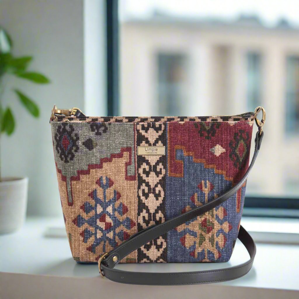 Canvas crossbody bag in vibrant Kilim geometric pattern with rich burgundy, navy, and taupe tones, featuring smooth black leather adjustable strap and polished brass hardware - front view by Umpie Handbags